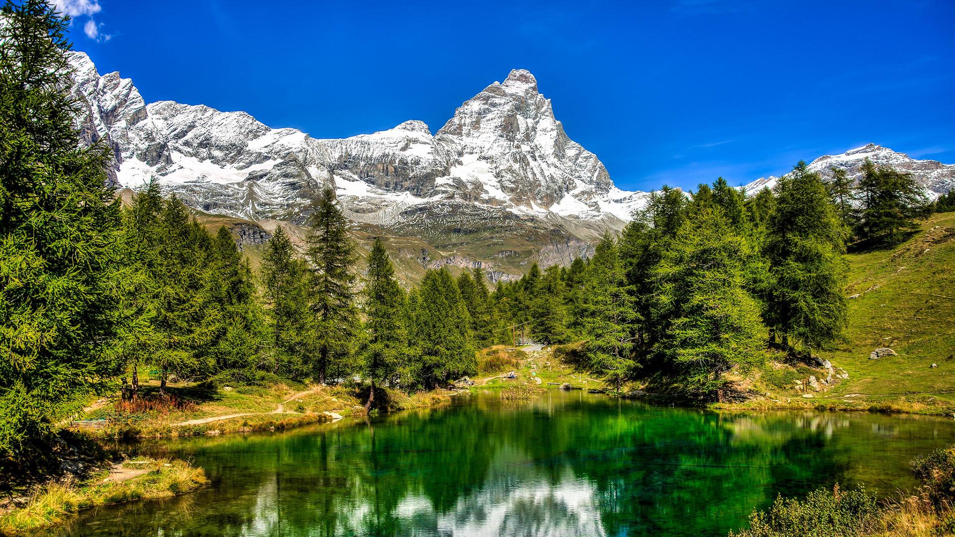 Lac alpin avec reflets montagneux, entouré d'arbres et de sommets enneigés.